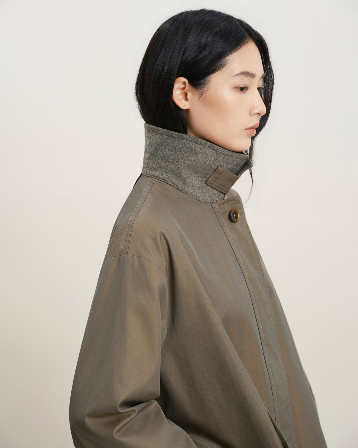 A model shown in profile wearing a high-neck, oversized olive-brown jacket with a contrasting textured gray collar, set against a plain light beige background.